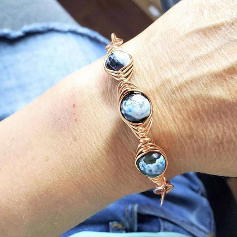 A close-up shows a wrist in blue jeans wearing the Alexa Martha Designs Herringbone Wire Wrap Child Abuse Prevention Half Cuff Ribbon Bracelet, an artisanal Southwestern piece with three round blue marbled stones.