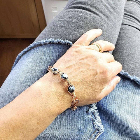 A person wearing grey jeans with frayed edges and a gold ring displays their wrist, adorned with the Alexa Martha Designs Herringbone Wire Wrap Child Abuse Prevention Half Cuff Ribbon Bracelet.
