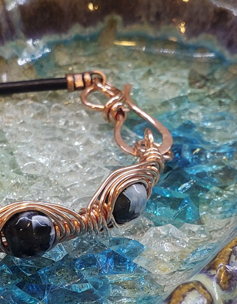 A close-up of the Herringbone Wire Wrap Child Abuse Prevention Half Cuff Ribbon Bracelet by Alexa Martha Designs, featuring black faceted beads and copper wire on a textured blue and clear glass surface.
