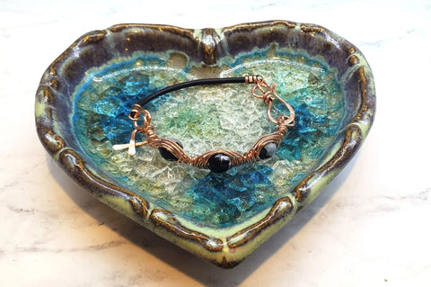 The Alexa Martha Designs Herringbone Wire Wrap Child Abuse Prevention Half Cuff Ribbon Bracelet lies on a blue and green heart-shaped ceramic dish filled with glass pieces.