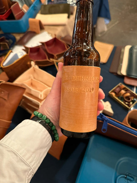 A hand holds a Sully + Proper Supply Handmade Leather Koozie: Can or Bottle, engraved with “THE DERRICKS 03/03/2019.” In the background, other handmade in Texas leather goods and accessories are displayed on a table.