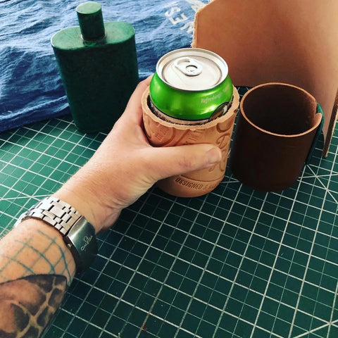 A tattooed hand wearing a wristwatch holds a can in the Sully + Proper Supply Handmade Leather Koozie on a green grid cutting mat. A green flask, blue cloth, and other leather items are also visible on the table.