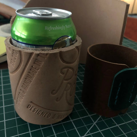 A green soda can is in a Sully + Proper Supply Handmade Leather Koozie (Can or Bottle) with embossed text, while an empty dark brown leather holder lies beside it on a green cutting mat.