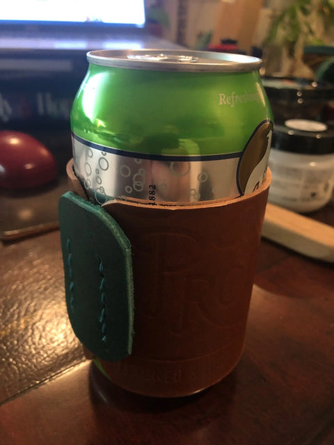 A green soda can sits in the Sully + Proper Supply Handmade Leather Koozie: Can or Bottle with turquoise stitching, on a wooden table with a keyboard and blurred background objects.