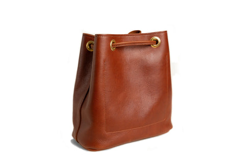 The Urban Southern Urban Bucket Bag, a brown leather handbag with gold eyelets and a drawstring closure, handcrafted in the USA, is shown on a white background.