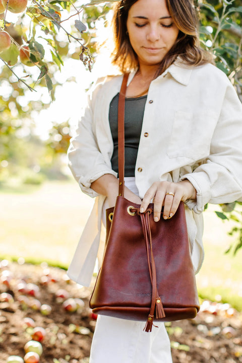 A woman stands outdoors by an apple tree, wearing a light jacket and holding the Urban Southern Urban Bucket Bag, handcrafted in the USA. Apples lie on the ground as she looks down with a gentle expression.