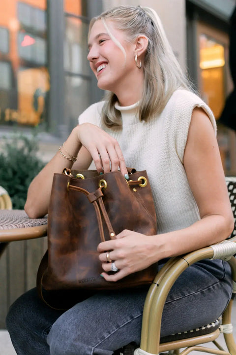 A smiling blonde woman sits at an outdoor café table, wearing a sleeveless cream top and gray jeans, holding the Urban Southern Urban Bucket Bag—handcrafted leather and made in the USA—on her lap.
