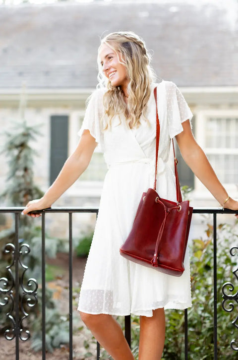 A smiling woman with wavy blonde hair stands outdoors by a black railing, wearing a white short-sleeve dress and carrying the Urban Southern Urban Bucket Bag. A house and greenery are softly blurred in the background.