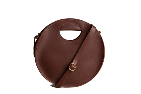 The Urban Southern Britt Circle Crossbody is a round, brown leather bag featuring an adjustable strap and cutout handle, shown on a white background.