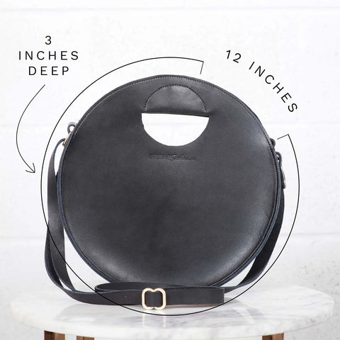 The Urban Southern Britt Circle Crossbody, a round black leather bag with a cut-out handle, adjustable shoulder strap, and gold buckle, sits on marble. Versatile as a crossbody, it measures 12 wide and 3 deep.