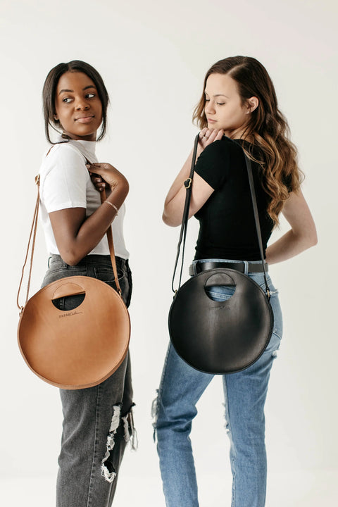 Two women stand back-to-back, each carrying an Urban Southern Britt Circle Crossbody—one in tan, one in black. Their casual outfits and the bags’ versatile design make a stylish statement against a plain white background.