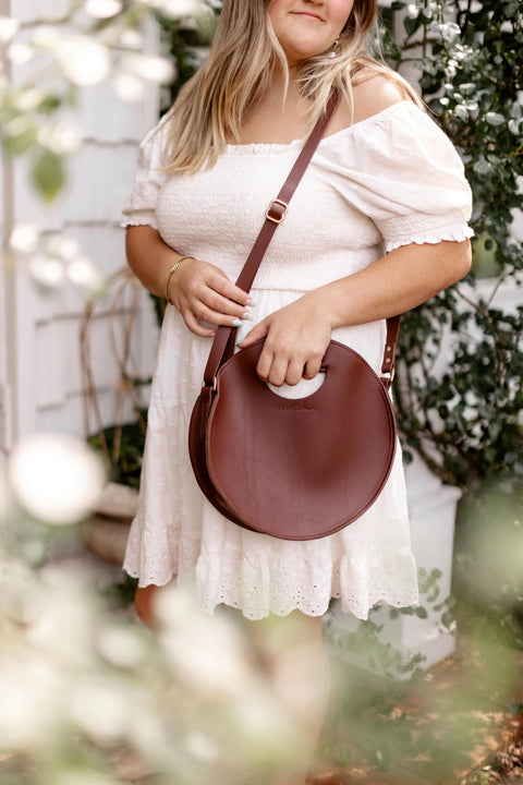 A woman outdoors, surrounded by greenery, wears a white off-shoulder dress and holds the Urban Southern Britt Circle Crossbody—a round brown leather bag with a versatile design. Her face is partially out of frame.