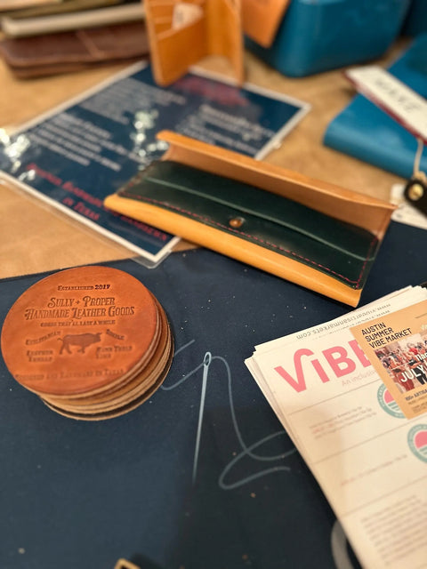 A set of customizable, personalizable Sully + Proper Supply leather coasters sits with a brown and green wallet, a menu, and a “Vibe” magazine on a table at a market or craft fair.