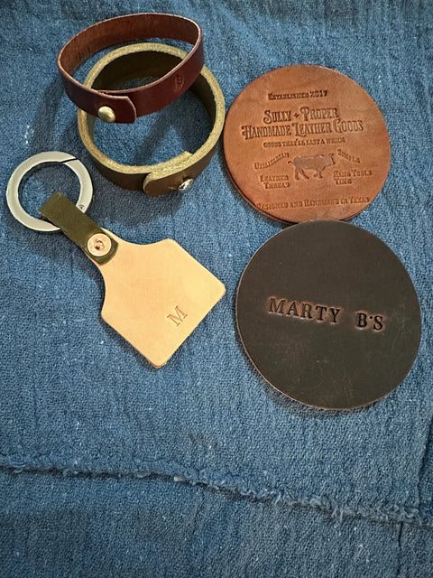 A set of handmade leather goods on blue fabric includes two bracelets, an M-initial keychain, and Sully + Proper Supply customizable coasters personalized with MARTY BS.