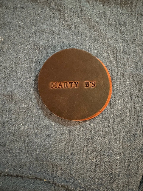 A round, brown handmade leather coaster from Sully + Proper Supply’s customizable Coasters line rests on a textured, dark blue fabric surface.