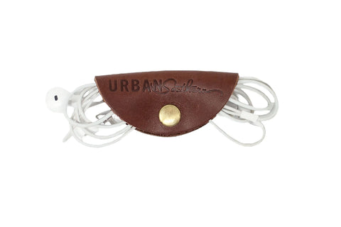 The Urban Cord Wrap by Urban Southern features brown leather and a brass snap button, embossed with the brand. It keeps white earphones neatly organized in style.