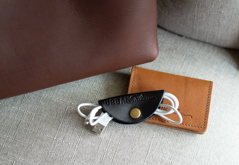 A white USB cable, neatly organized with the Urban Southern Urban Cord Wrap in black leather, sits beside a tan leather cardholder on a light fabric surface near a brown leather bag.