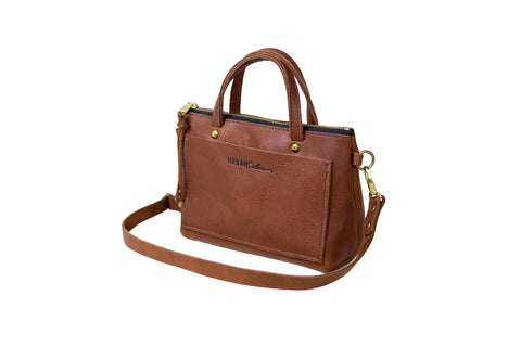 The Cassidy Crossbody by Urban Southern is a brown leather handbag with two short handles, gold hardware, an outer pocket, and a detachable strap. Its compact design is shown against a white background.