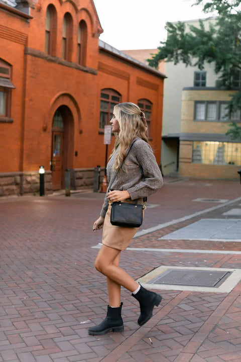 A woman with long, wavy hair walks on a city sidewalk in a brown sweater, tan skirt, black boots, and carries the Cassidy Crossbody by Urban Southern. Brick buildings and trees line the background.