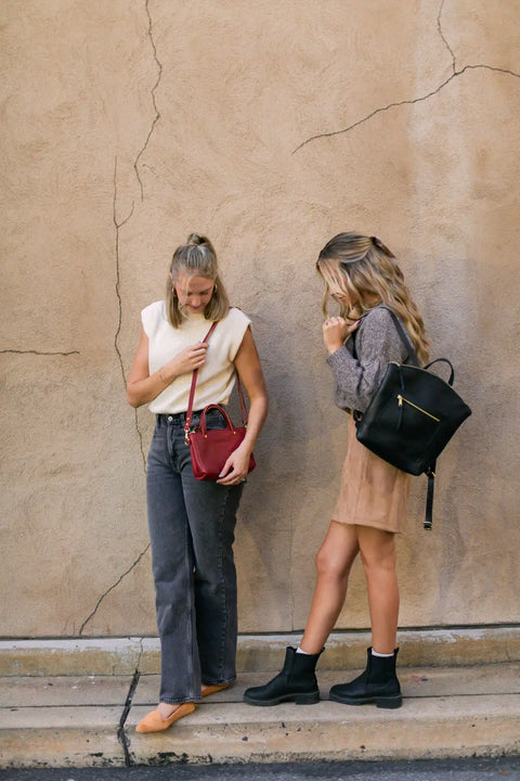 Two women stand against a textured beige wall. One wears a sleeveless top, gray jeans, and carries an Urban Southern Cassidy Crossbody in red. The other wears a gray sweater, tan skirt, black boots, and carries a black backpack. Both look downwards.