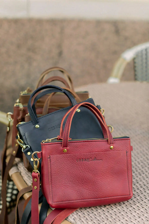 Three compact Cassidy Crossbody leather handbags from Urban Southern in red, navy blue, and brown are displayed on a woven surface, each with gold hardware, a stylish leather strap, and the Urban Southern name embossed on the front.
