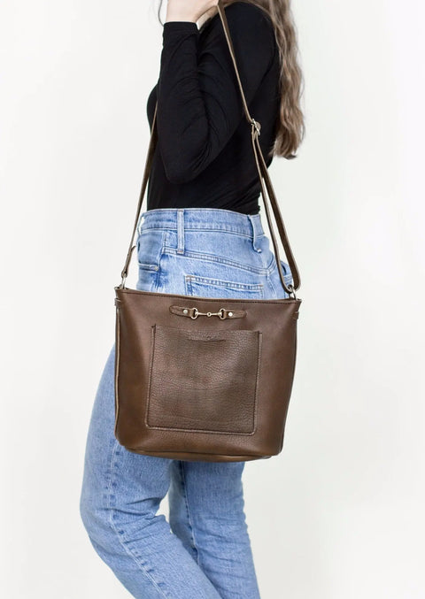 A woman in a black long-sleeve top and blue jeans carries The Lexington Julep by Urban Southern, an American-made leather crossbody with a front pocket and gold hardware, photographed against a white background.