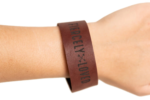 The Urban Southern Fiercely Loved Cuff, a brown full-grain leather bracelet with FIERCELY LOVED in black letters, is shown on a wrist against a white background.