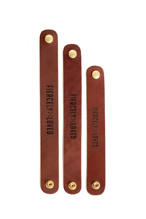 Three Urban Southern Fiercely Loved Cuff bracelets in brown full-grain leather, each a different length, feature gold snaps and FIERCE + LOVED stamping. All are handcrafted in the USA and displayed vertically on a white background.