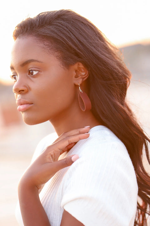 A woman with long dark hair wearing a white top and Urban Southern’s Everyday Earrings with bronze-plated hooks gazes thoughtfully to the side, her hand near her neck against a softly blurred outdoor background.
