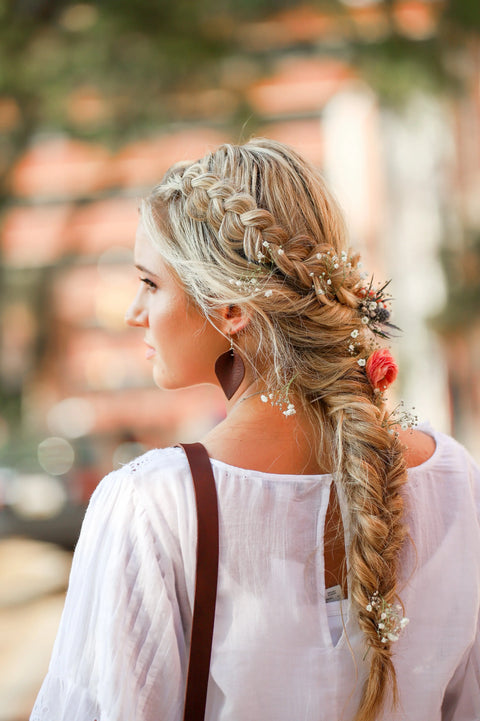 A woman with blonde hair in a loose braid decorated with small flowers, wearing a white blouse and Urban Southerns Everyday Earrings with bronze-plated hooks, is seen from behind outdoors.