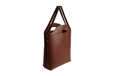 The Urban Southern Market Tote is a brown leather bag featuring dual handles and a cut-out hand grip, shown standing upright against a white background.