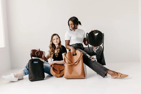 Two women sit on the floor against a white wall, showing off Urban Southern handbags in black, brown, and tan—including the versatile Market Tote, ideal as a reusable grocery bag. Both wear jeans and neutral tops for a casual style.