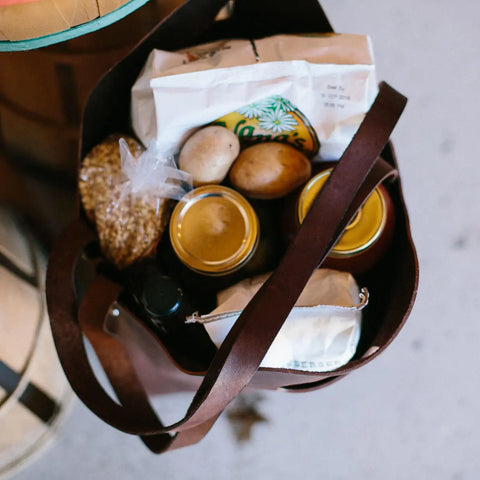 The Urban Southern Market Tote, a reusable grocery bag, holds groceries like two glass jars, potatoes, grains in a plastic bag, a bottle, and paper-wrapped items as it rests on a light surface.
