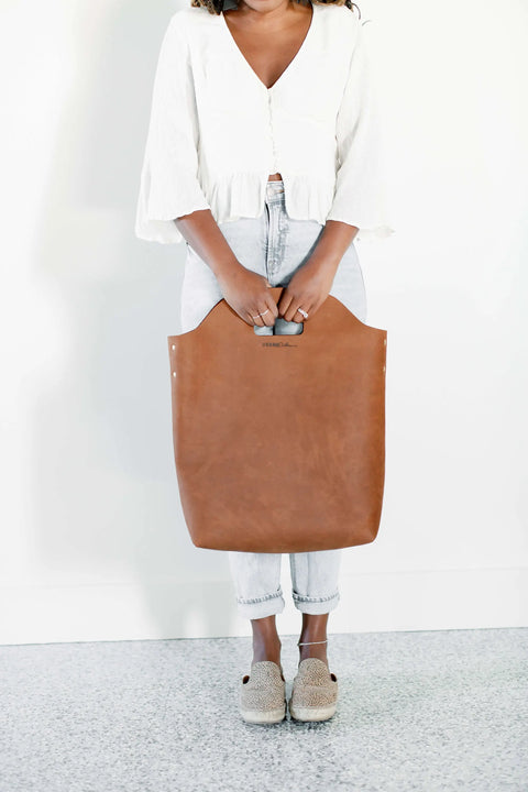 A woman in a white blouse and light jeans holds the Urban Southern Market Tote, a spacious and versatile bag for any outing. She stands against a white wall wearing beige woven slip-on shoes; her face is not visible.