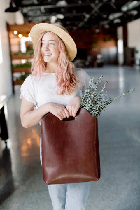 A young woman with pink hair, in a straw hat and white shirt, smiles while holding the Urban Southern Market Tote filled with eucalyptus branches, standing in a bright, modern indoor space.