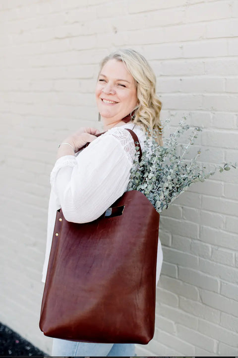 A smiling blonde woman in a white blouse stands before a light brick wall, carrying the Urban Southern Market Tote over her shoulder, filled with sprigs of greenery.