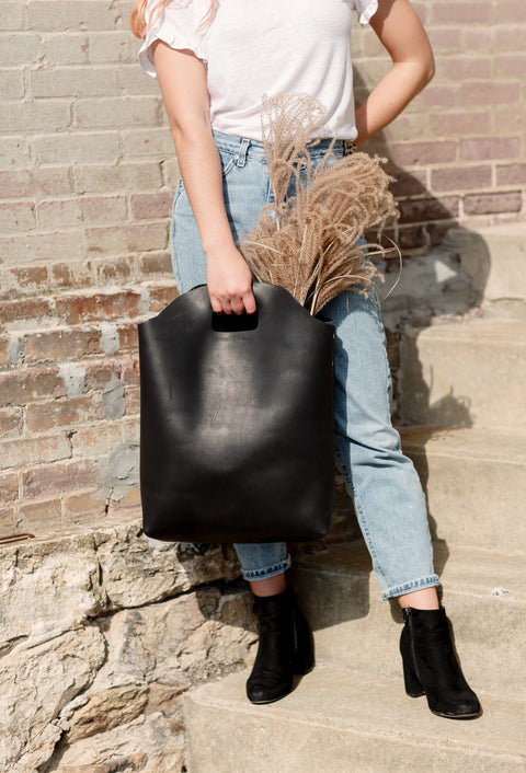 A person in a white T-shirt, light blue jeans, and black boots stands on outdoor steps with the Urban Southern Market Tote filled with decorative dried grass. A weathered brick wall provides the background.