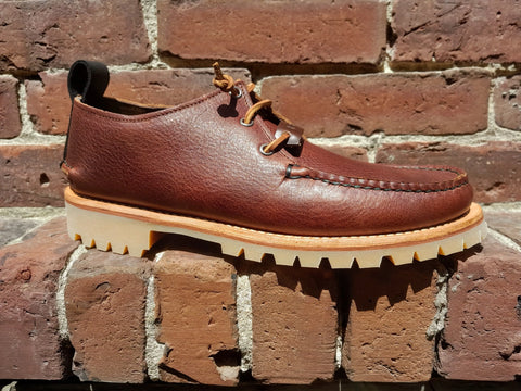 The BBH Kineo, a handcrafted moccasin in brown premium leather with tan rugged soles, is showcased against a red brick background bathed in bright sunlight.