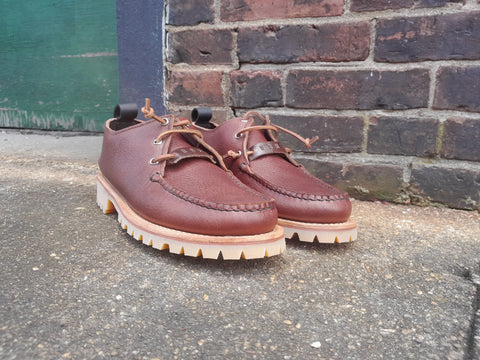 A pair of BBH Kineo handcrafted moccasin-style brown leather shoes with rugged tan soles are displayed outdoors on concrete near a brick wall and green wooden door.