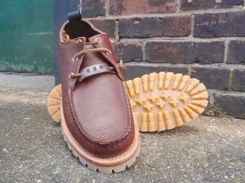 The BBH Kineo is a handcrafted brown premium leather moccasin with tan laces, shown outdoors—one upright and one on its side to highlight its chunky, rugged sole with deep treads for durable, sustainable wear.