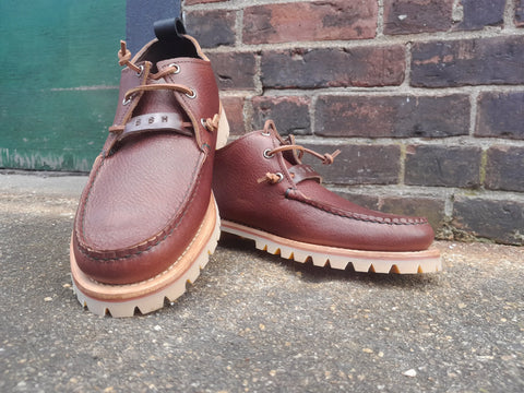 The BBH Kineo, handcrafted moccasin-style shoes made from premium leather with thick, ridged tan soles, are displayed outdoors on a concrete surface against a brick wall.