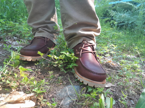 A person wearing BBH Kineo handcrafted brown leather moccasins and khaki pants stands on a grassy, sunlit forest floor with plants and rocks around their feet.