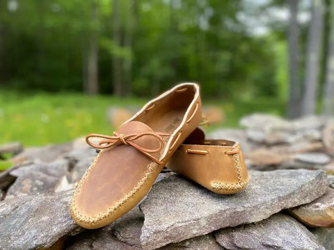 The BBH Lone Sole Old Town Mocc, a pair of brown handcrafted moccasins with stitched edges and laces, is displayed outdoors on flat rocks with a blurred green forest in the background.