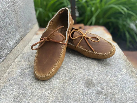 The BBH Lone Sole Old Town Mocc, a pair of brown handcrafted moccasins with visible stitching and leather laces, is shown outdoors on stone with green plants in the background.