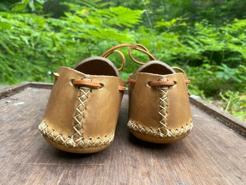 The BBH Lone Sole Old Town Mocc moccasins, featuring visible stitching, are displayed on a wooden surface outdoors amid green foliage, highlighting these timeless handcrafted shoes.