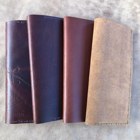 Four handmade Rancher Log Covers by Sully + Proper Supply, in rich brown and tan shades, are vertically arranged on a textured beige surface. Subtle stitching and embossed designs showcase timeless American craftsmanship.