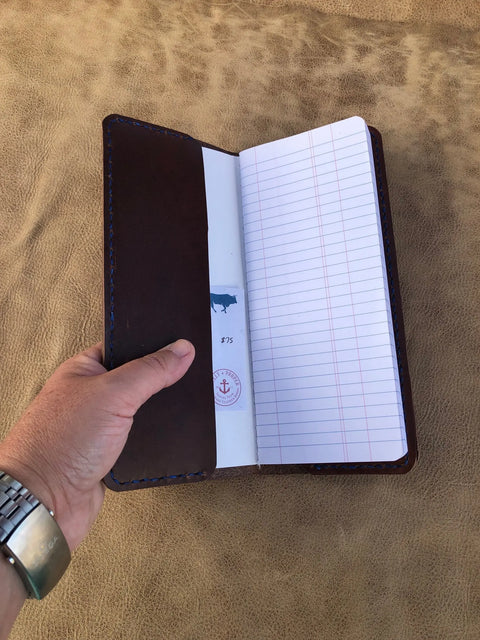 A hand holds an open Sully + Proper Supply Rancher Log Cover with lined pages. The inside cover features a blue animal silhouette and red anchor stamp, set against a textured tan background.