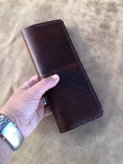 A hand holds a dark brown Sully + Proper Supply Rancher Log Cover with blue stitching against a light brown textured background. The person wears a silver watch and ring—ideal for fans of rancher journal style or leather log covers.