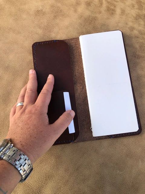 A hand with a silver watch and ring opens a Sully + Proper Supply Rancher Log Cover, made in the USA, to reveal a white notebook and card on a tan textured surface.