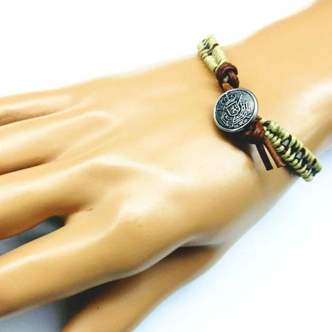 A mannequin hand displays the Alexa Martha Designs Leather Seed Bead Rattlesnake Tail Weave Bracelet, handcrafted with green and brown cords and accented by a round metallic Tree of Life charm engraved with a design.